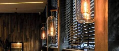 L’intérieur d’un restaurant présente des murs en bois, des stores métalliques et des lampes suspendues. Les tables sont dressées avec des couverts et des serviettes.