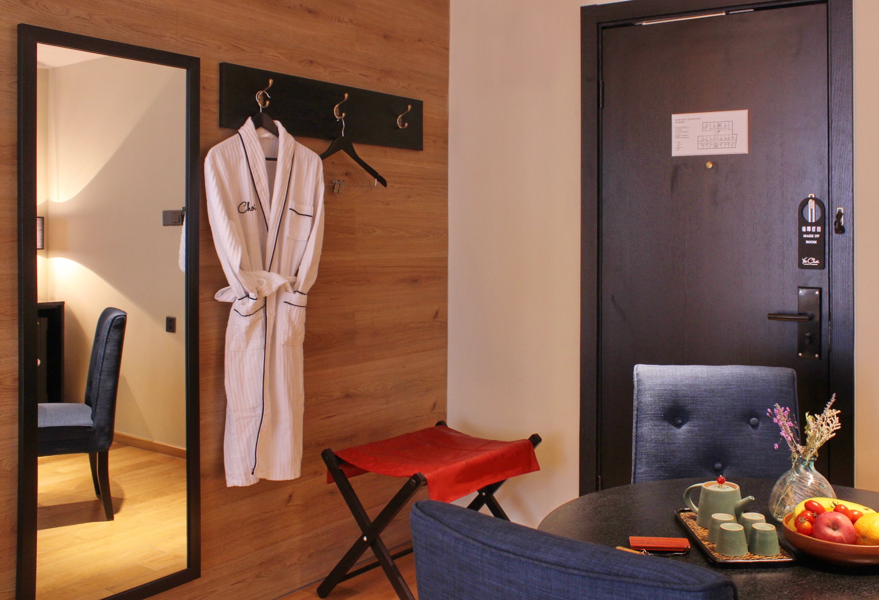 A hotel room with a wooden wall, a robe hanging on a rack, a small red stool, a table with a vase and fruit, and a door with a keycard lock.