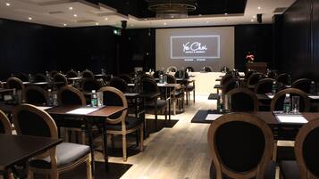 A conference room with rows of tables and chairs, a large screen displaying 'You Choi Hotels & Resorts', and a podium with flowers.