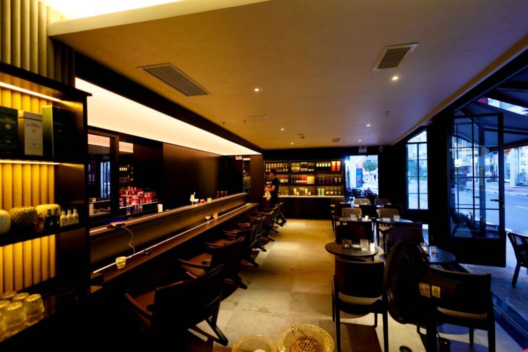 A bar area with a long counter, chairs, and shelves with bottles. There are tables and chairs in the foreground, and a view of the street through large windows.