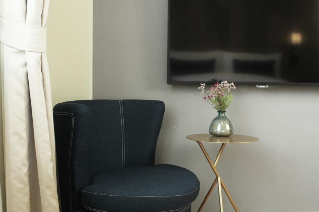 Una habitación con una silla tapizada de azul oscuro, una mesita redonda con un florero y un televisor de pantalla plana montado en la pared.