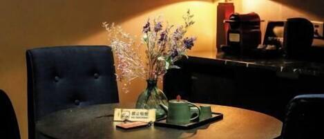 A dining area with a round table, two chairs, a vase with flowers, a book, and a coffee machine in the background.