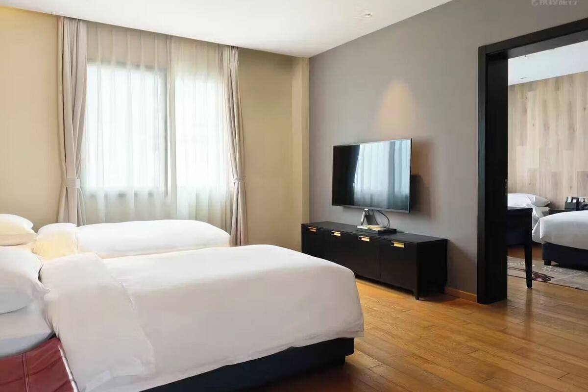 Une chambre d’hôtel moderne, dotée de deux lits, d’un téléviseur à écran plat, d’une commode en bois foncé et d’une grande fenêtre agrémentée de rideaux légers.