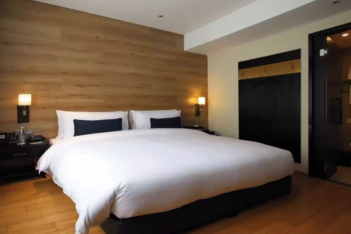 Une chambre d’hôtel moderne avec un grand lit, une tête de lit en bois, des lampes de chevet et une porte sombre.
