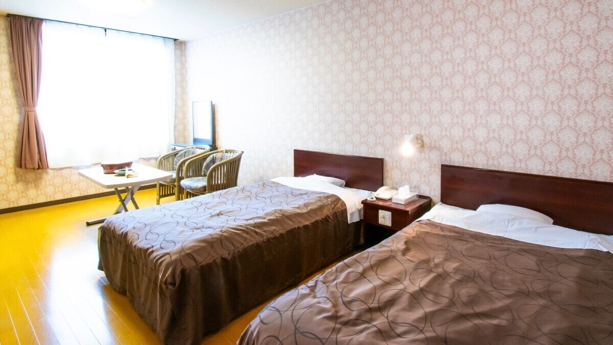 Une chambre d’hôtel avec deux lits, un bureau avec des chaises, une fenêtre avec des rideaux et un papier peint à motifs.