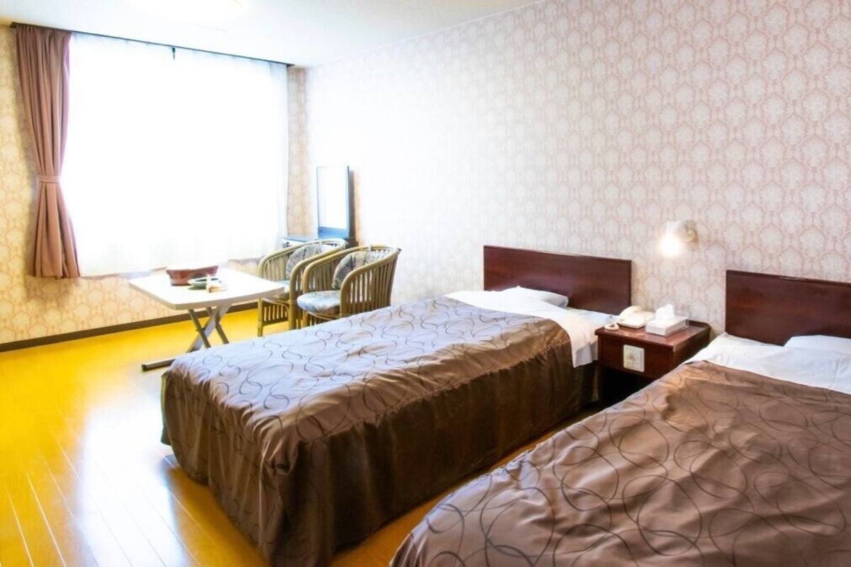 Ein Hotelzimmer mit zwei Betten, einem Schreibtisch mit Stühlen, einem Fenster mit Vorhängen und einem Wandpapier mit Muster.