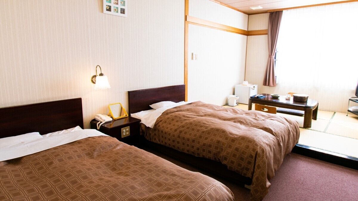 Quarto de hotel com duas camas, uma mesa de cabeceira com abajur, uma mesinha com uma cadeira e uma janela com cortina.
