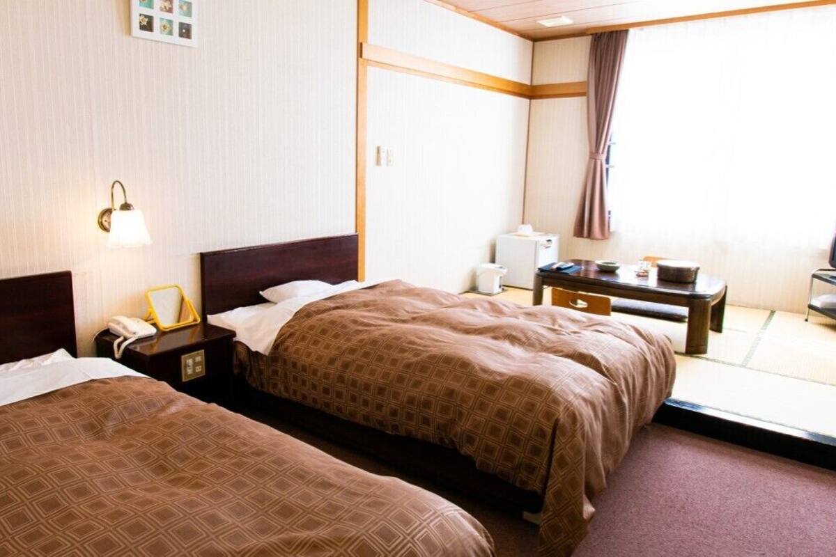 Ein Hotelzimmer mit zwei Betten, einem Nachttisch mit Lampe, einem kleinen Tisch mit Stuhl und einem Fenster mit Vorhang.