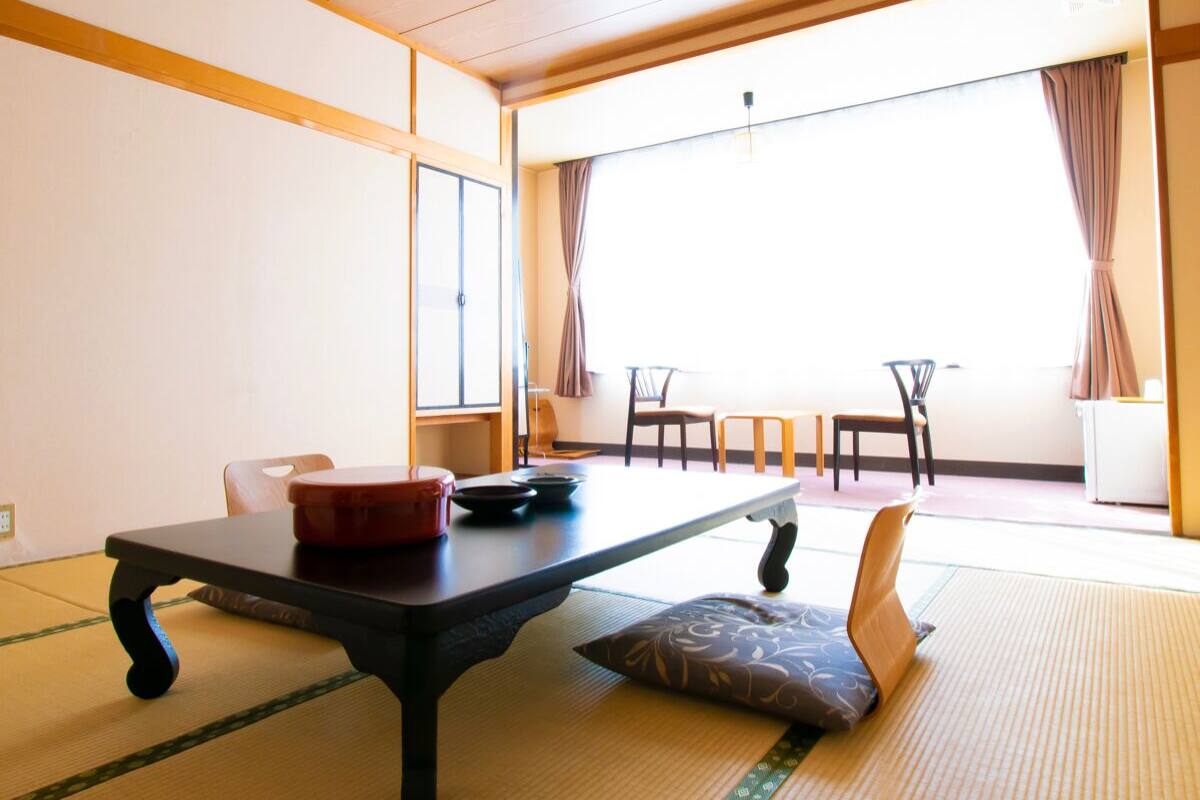Ein traditioneller japanischer Raum mit Tatami-Böden, einem niedrigen Tisch mit einem roten Behälter und Blick ins Freie durch große Fenster.