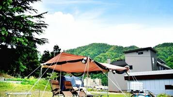 A camping setup with a tent, chairs, and a table in a grassy area with a house and hills in the background.