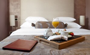 Una cama bien hecha con sábanas y almohadas blancas, una mesita de noche con una bandeja que contiene una tetera, tazas, un vaso de zumo de naranja y algunos alimentos.