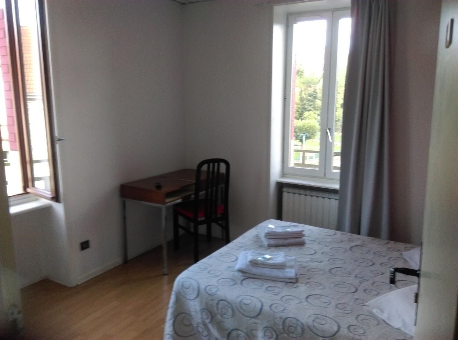 A small, well-lit room with a single bed, a desk, and a chair. There is a window providing natural light and a view of greenery outside.
