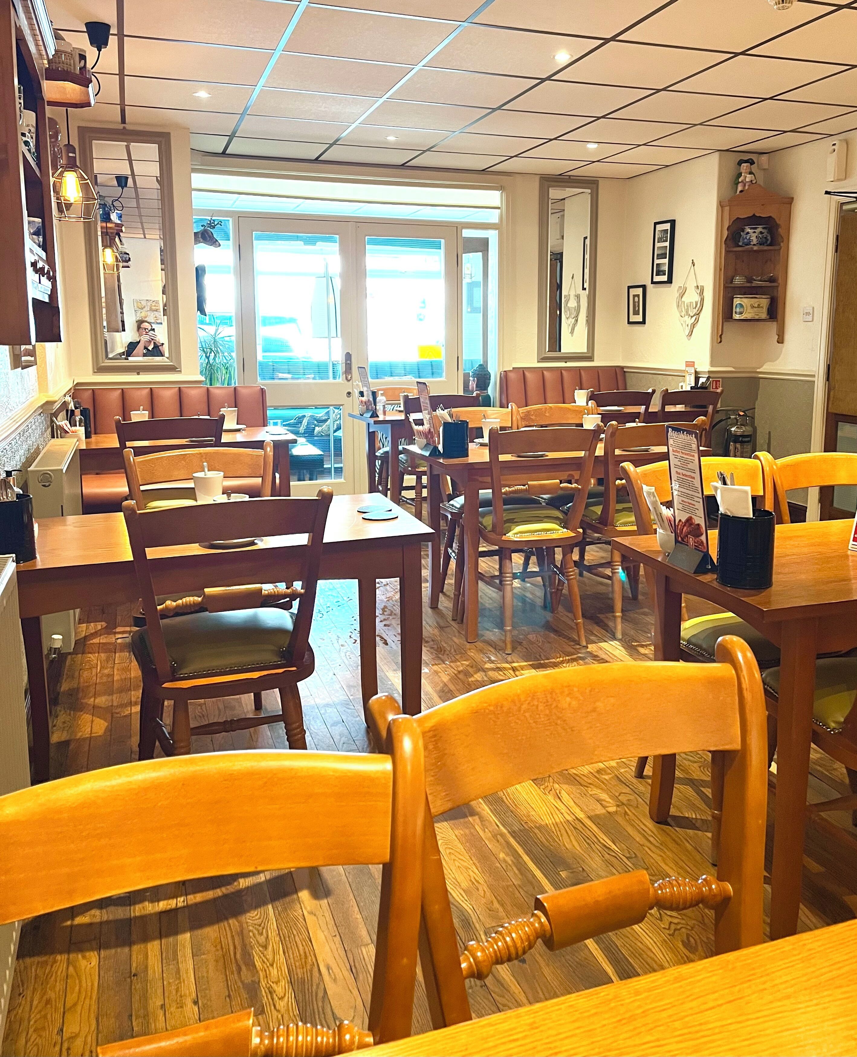 A restaurant interior with wooden tables and chairs, a tiled ceiling, and large windows allowing natural light.