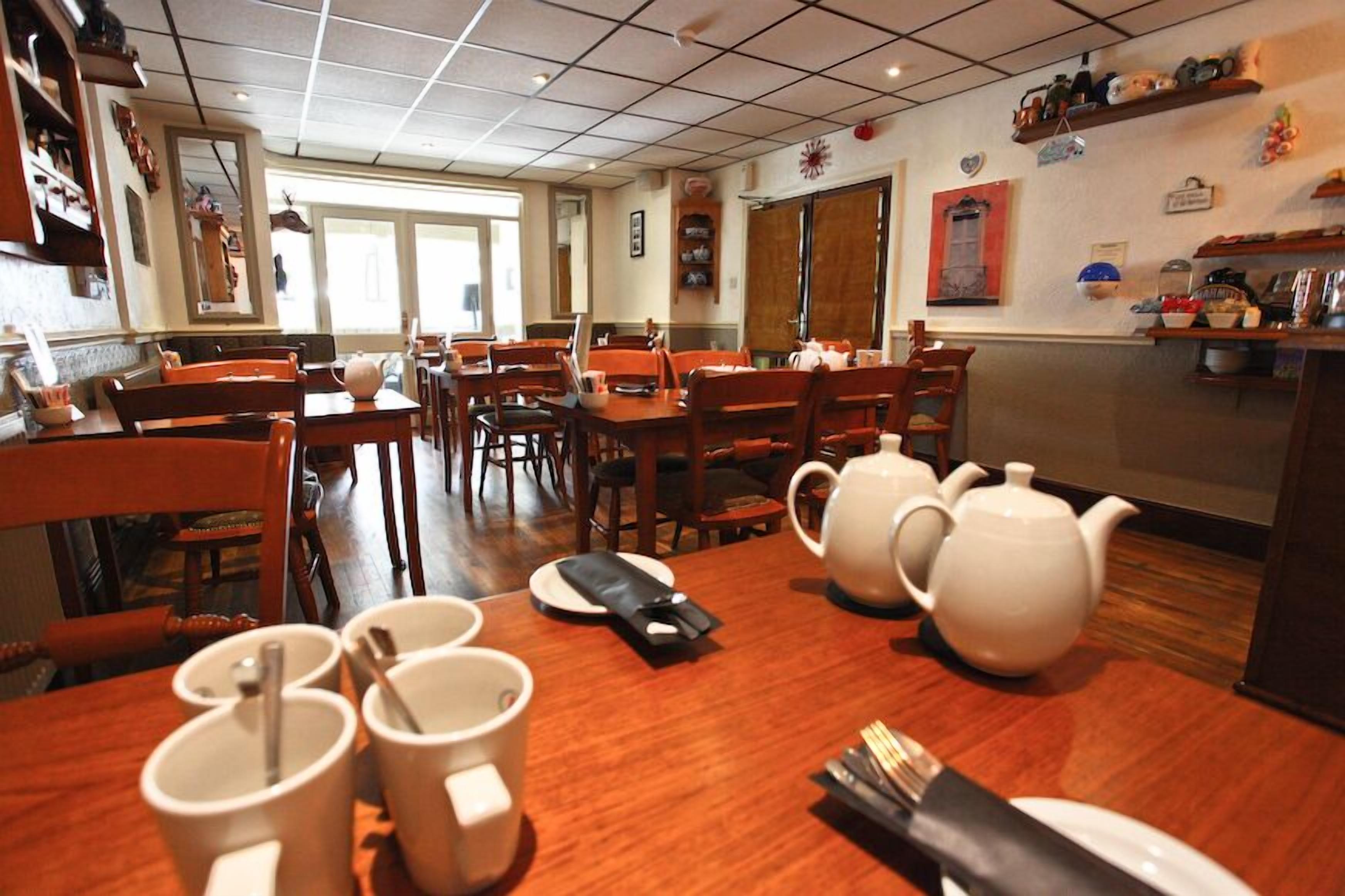 A dining area with wooden tables and chairs, white teapots, and a kitchen counter with various items.