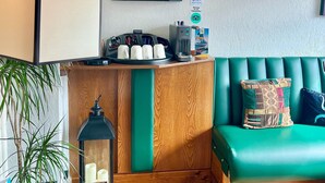 Un área de recepción con un sofá verde, una lámpara, una estatua de Buda, un estante con objetos y un cesto de basura.