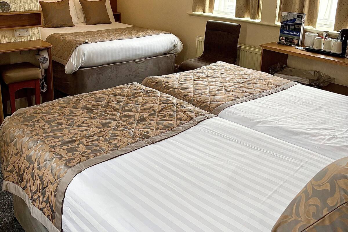 A hotel room with two beds, a desk, a chair, a window with curtains, a painting on the wall, and a ceiling with a smoke detector.