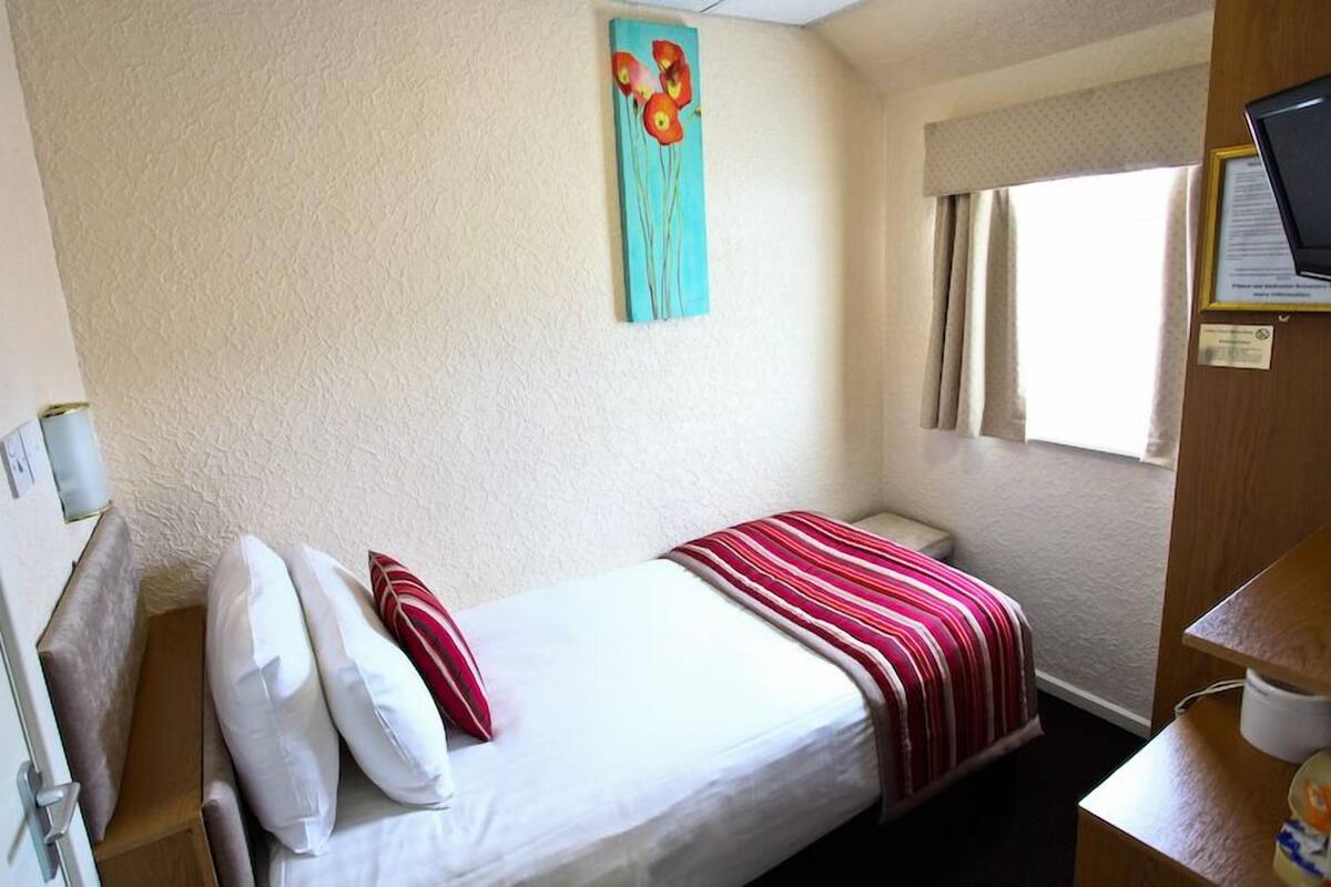 A single bed with white and red bedding, a small bedside table, a window with curtains, and a painting on the wall.