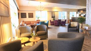 A spacious living area with multiple seating options, including sofas and armchairs, arranged around a central coffee table. There is a chandelier hanging from the ceiling, and the room is well-lit with natural light from large windows. - Bardufoss Hotell (Målselv)