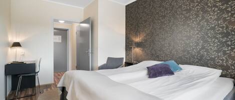 A hotel room with a large bed, a desk with a chair, a small rug, and a door leading to another room.