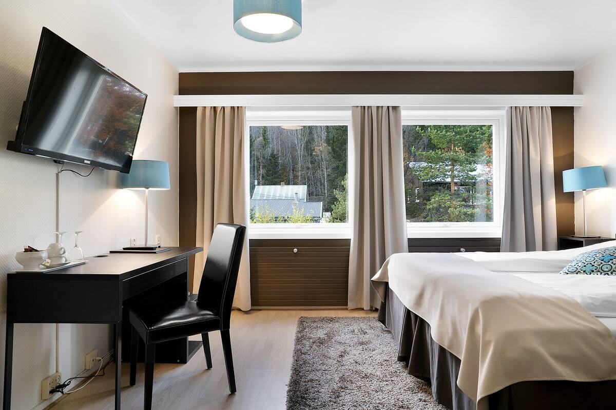 Habitación de hotel con cama, escritorio, silla, televisor y ventana grande con cortinas.