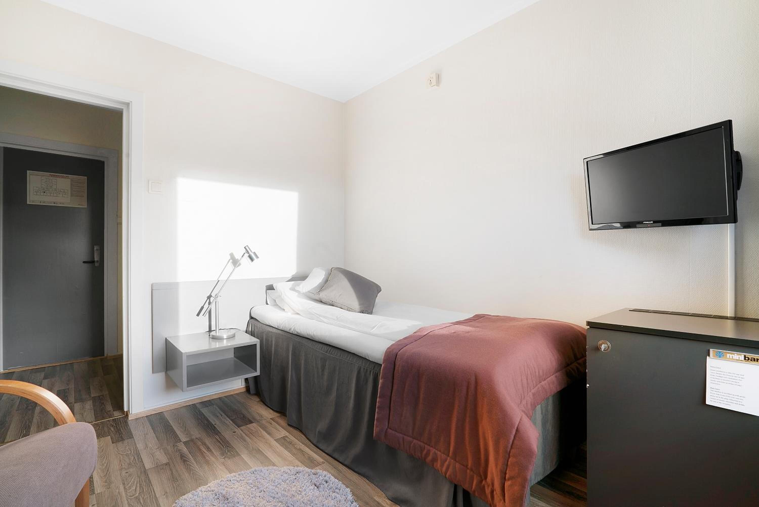 A small, modern room with a bed, bedside table, lamp, and a TV mounted on the wall.
