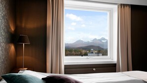 A hotel room with a bed, a nightstand, a lamp, and a window with a mountain view. - Bardufoss Hotell (Målselv)