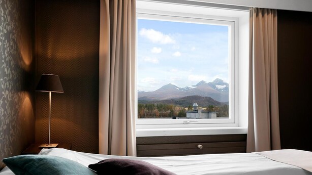 A hotel room with a bed, a nightstand, a lamp, and a window with a mountain view. - Bardufoss Hotell (Målselv)