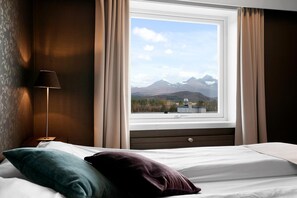 A hotel room with a bed, a nightstand, a lamp, and a window with a mountain view. - Bardufoss Hotell (Målselv)