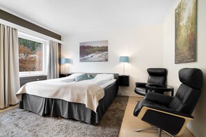 Ett modernt hotellrum med en säng, ett skrivbord, en stol och ett stort fönster med gardiner.