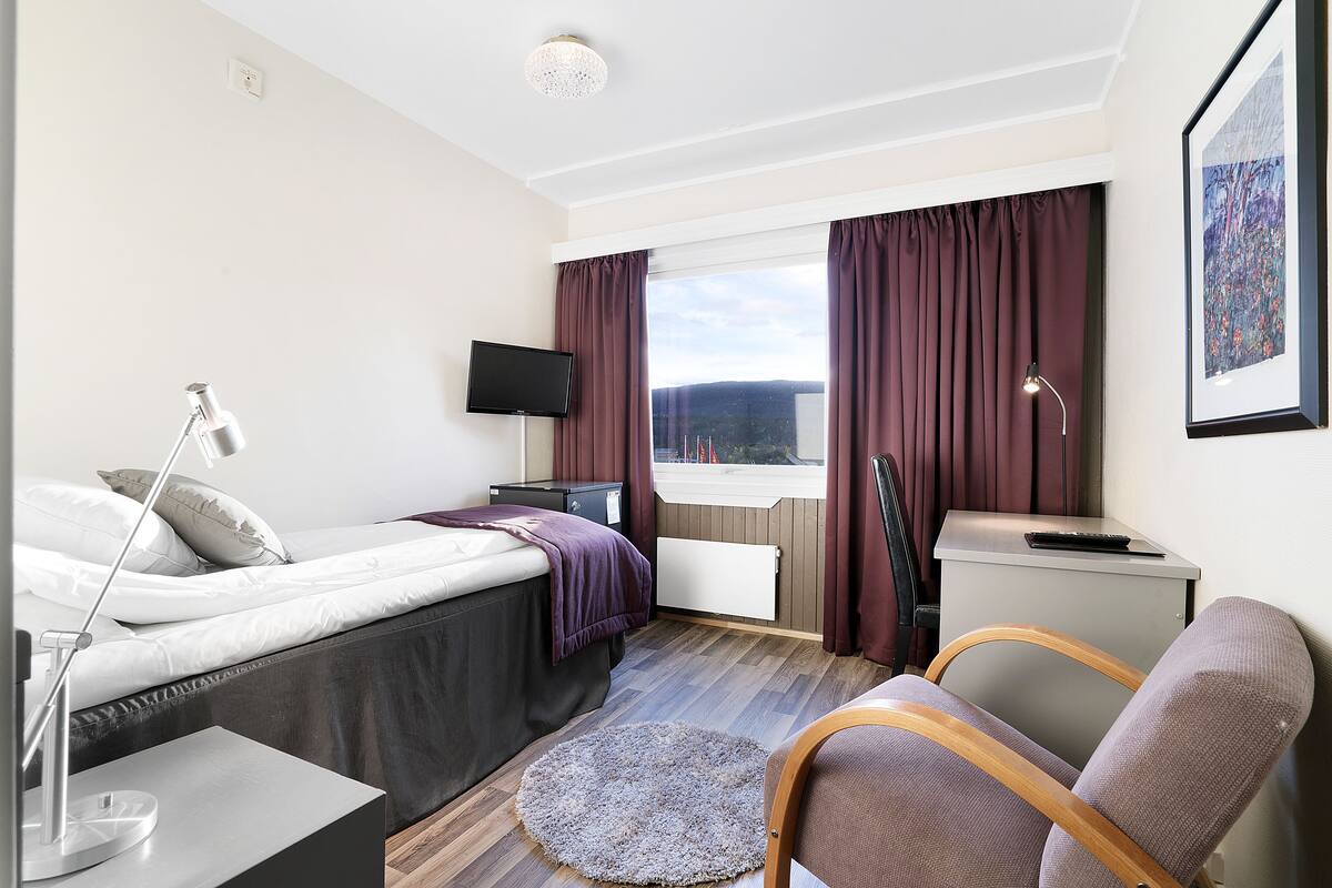 Una habitación de hotel moderna con cama, mesita de noche, lámpara, silla, escritorio y ventana con cortinas.