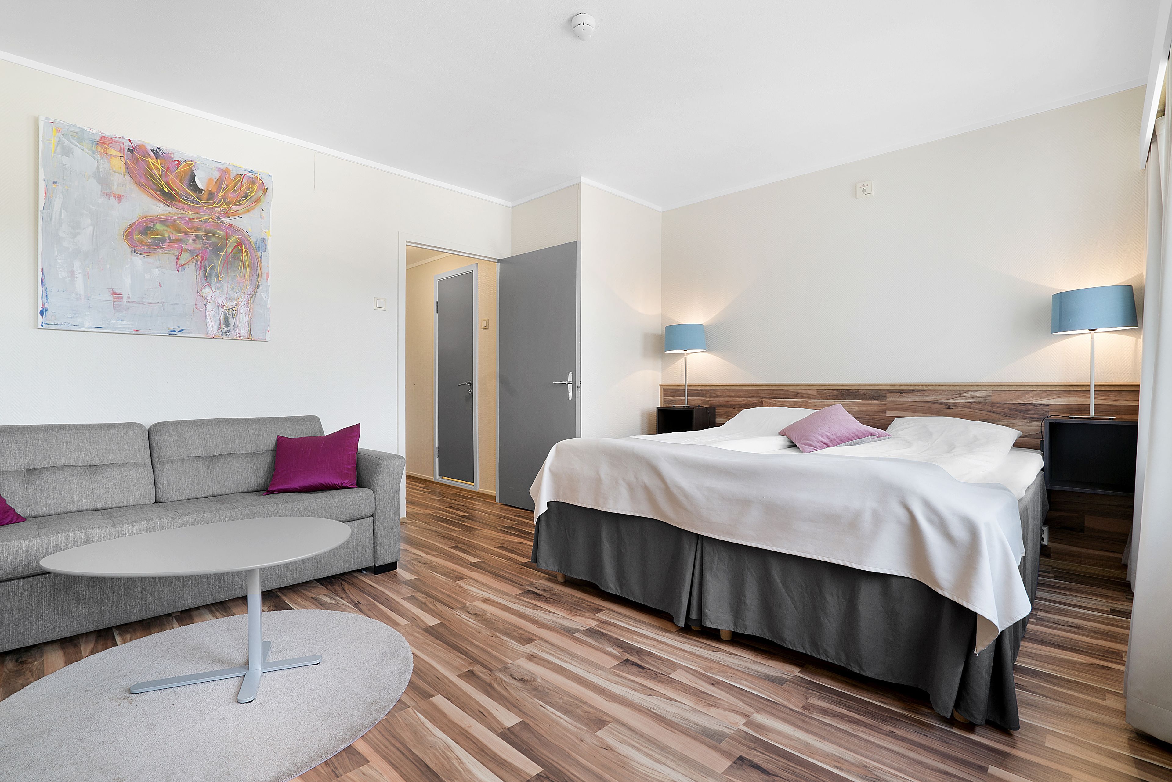 Una habitación de hotel moderna con un sofá gris, una mesa redonda, una cama con falda gris y un cuadro abstracto grande en la pared.