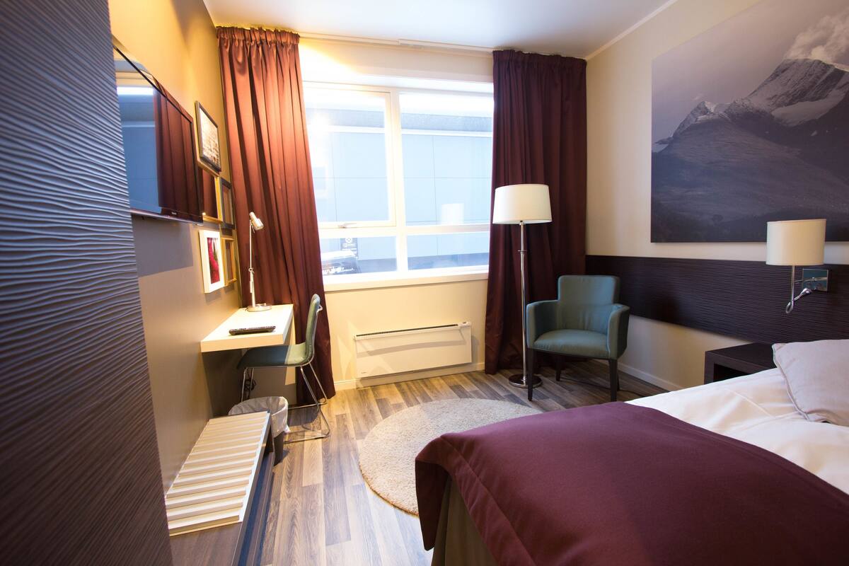 Habitación de hotel con cama, escritorio, silla y un amplio ventanal con cortinas.
