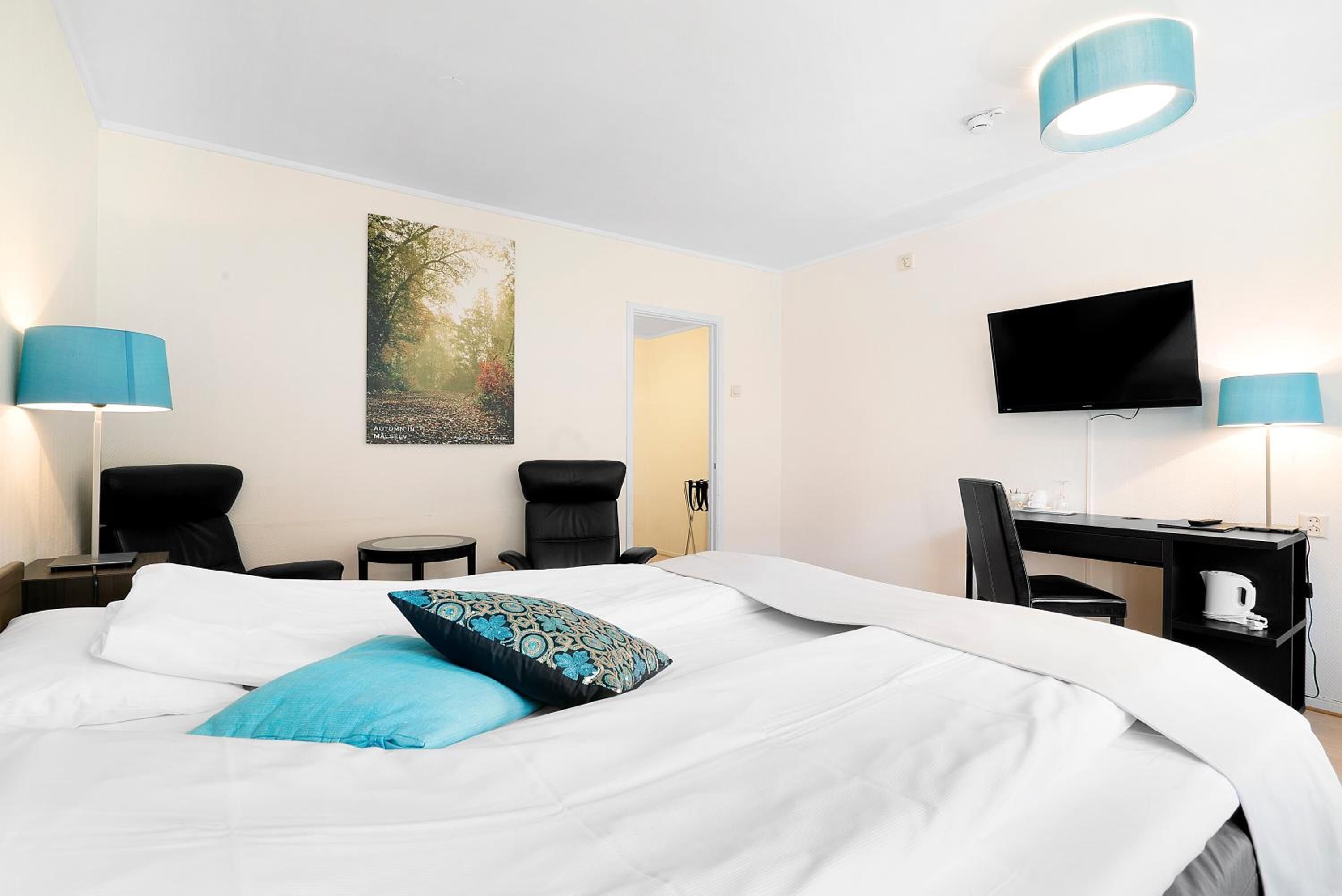 Quarto de hotel com uma cama, duas cadeiras, uma escrivaninha com uma TV e um quadro na parede.