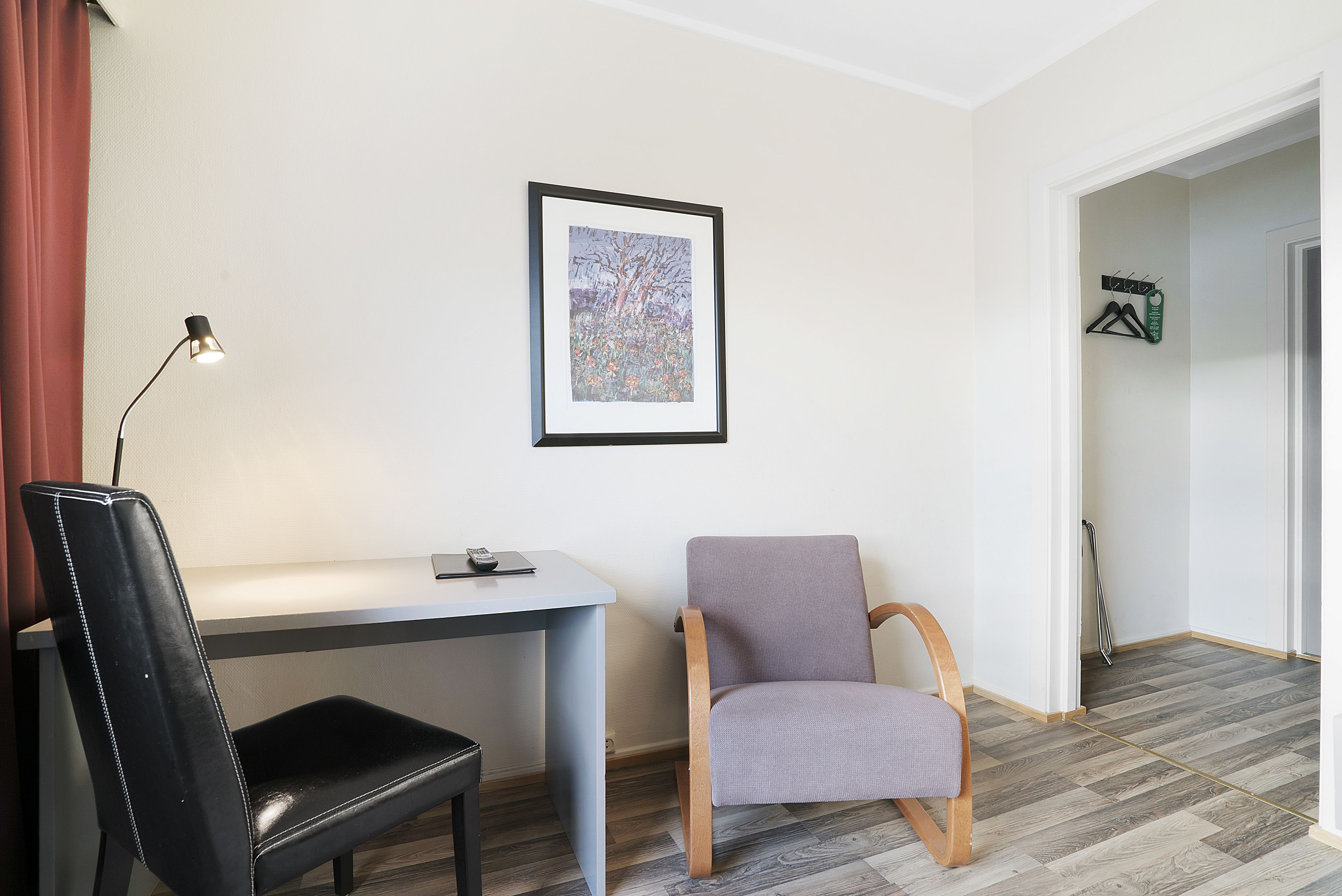 Una habitación con escritorio, una silla de cuero negro, una butaca gris acolchada, un cuadro en la pared y un perchero con ganchos.