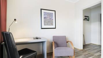Una habitación con escritorio, una silla de cuero negro, una butaca gris acolchada, un cuadro en la pared y un perchero con ganchos.