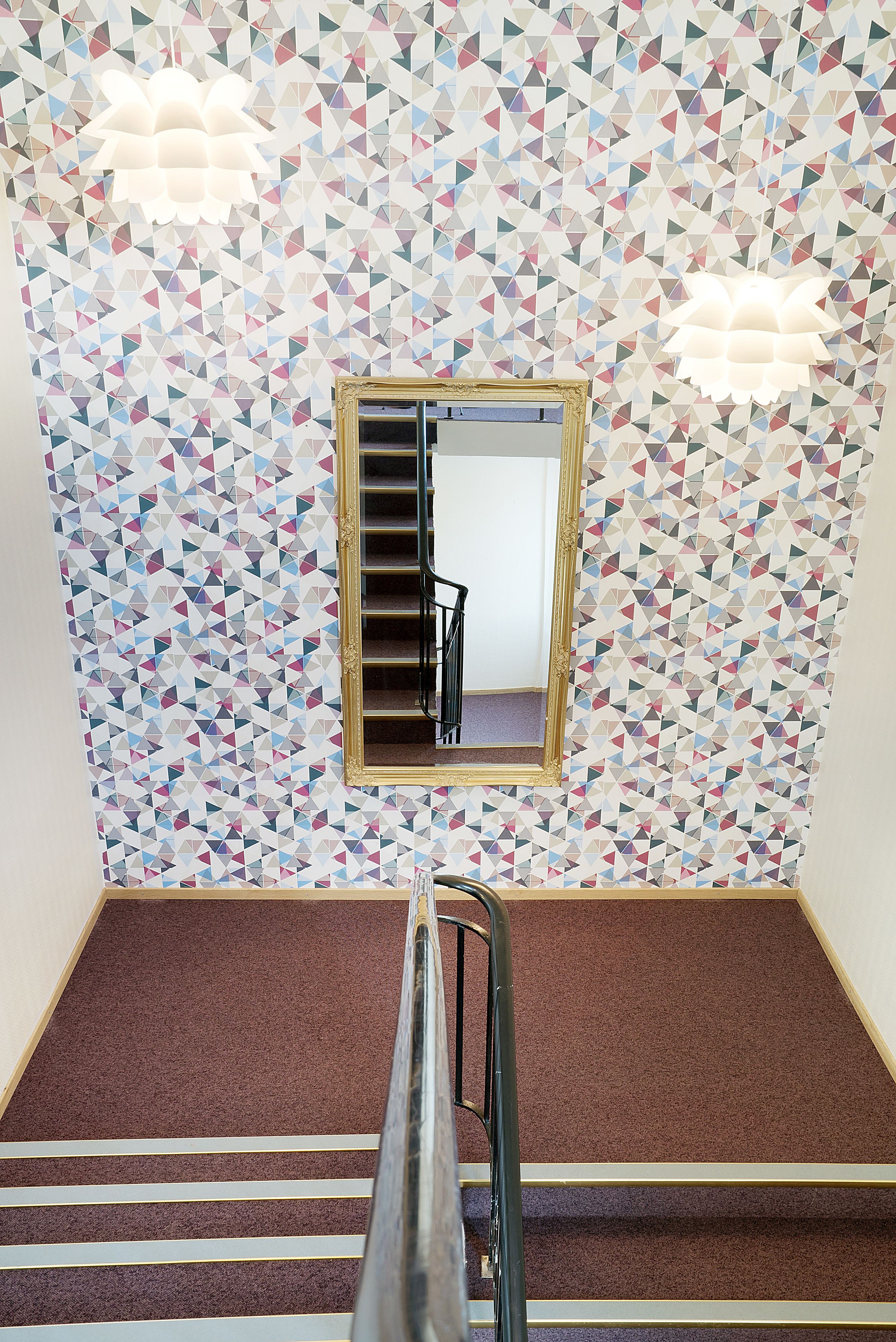 A room with a geometric patterned wall, a mirror with a gold frame, and a brown carpeted floor.