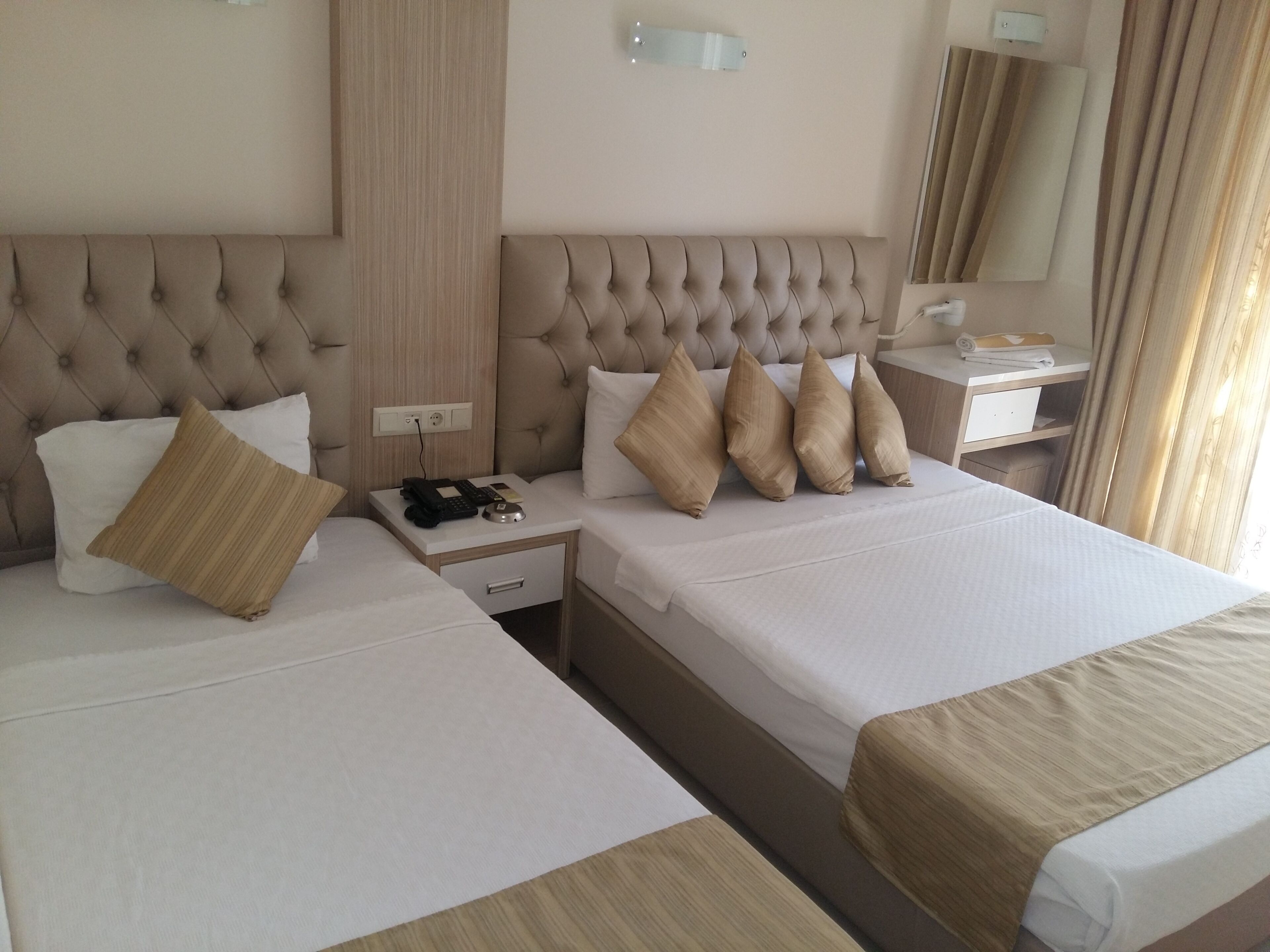 A hotel room with two beds, a bedside table with a phone and remote, a nightstand, a mirror, and a window with curtains.