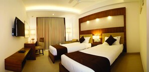 A hotel room with two beds, a desk, a chair, a TV, and a large window with curtains. - Urvashi Residency (Chennai)