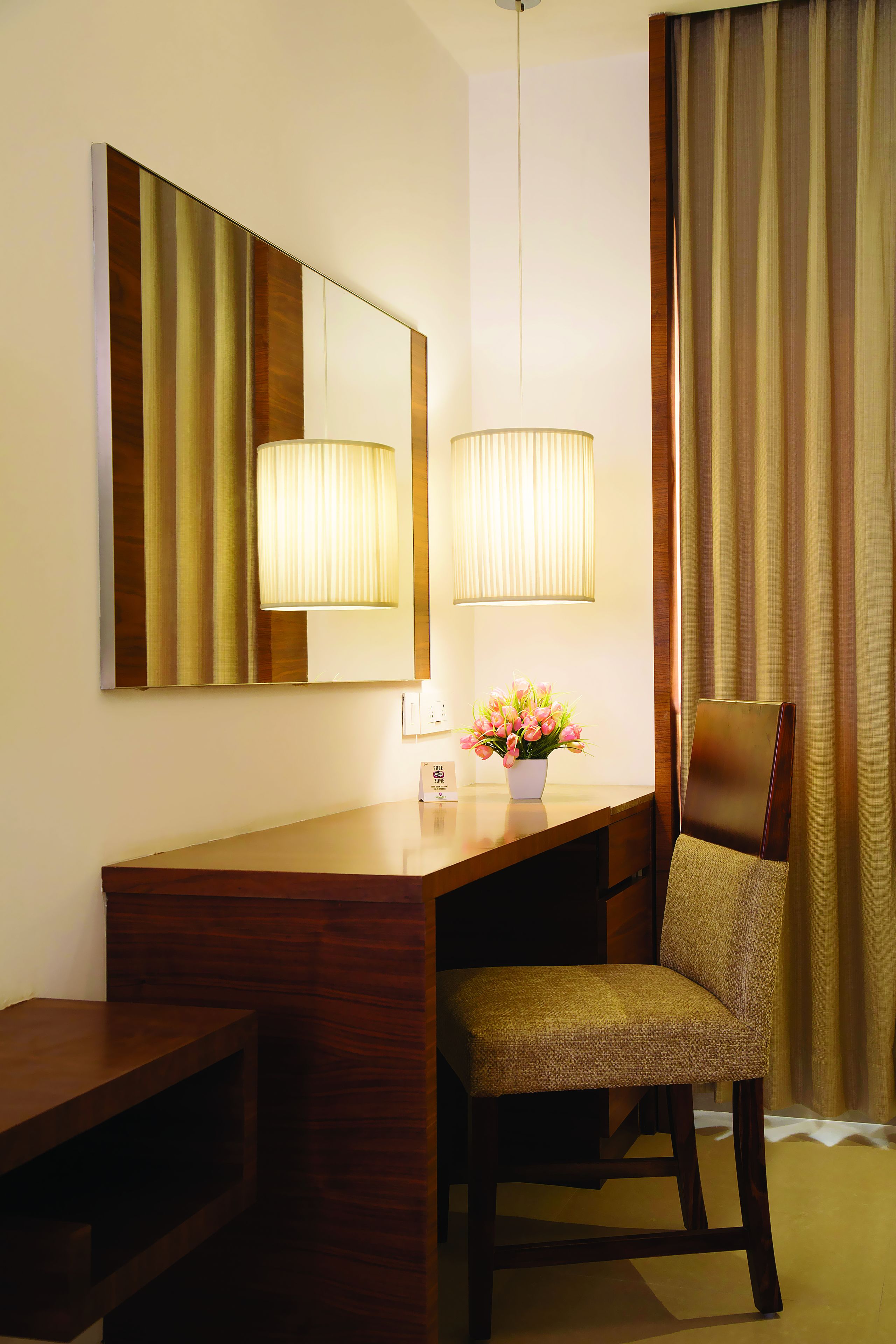 A hotel room with a wooden desk, a chair, a large mirror, a lamp, and a vase with flowers.