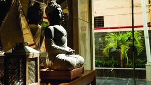 Une statue représentant une figure assise, placée sur une plateforme en bois, avec une lanterne ajourée à gauche et une vue sur des plantes d’intérieur à travers une fenêtre en verre.