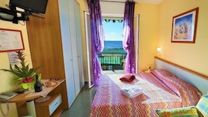 A hotel room with a bed, a sofa, a desk with a computer, a plant, and a view of the sea through the window. - Hotel Lucerna (Taggia)