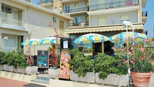 A seaside restaurant with outdoor seating, a menu board, and potted plants. - Hotel Lucerna (Taggia)
