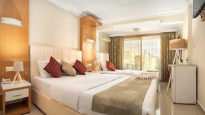 Ein Hotelzimmer mit zwei Betten, einem Nachttisch mit Lampe, einem Kühlschrank und einem großen Fenster mit Vorhängen.