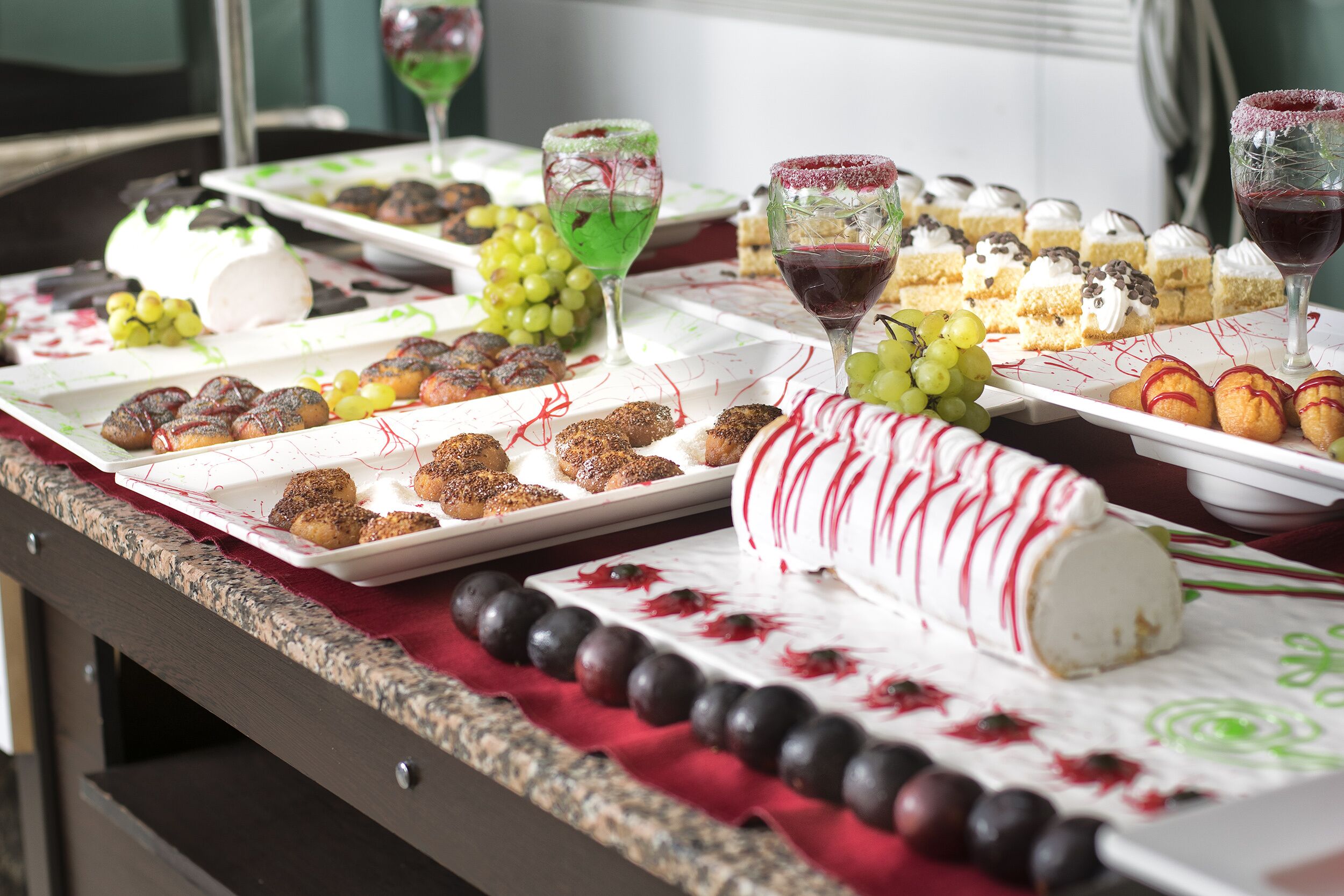 A buffet table with various desserts and fruits, including a large white cake with red drips, and several glasses of wine.