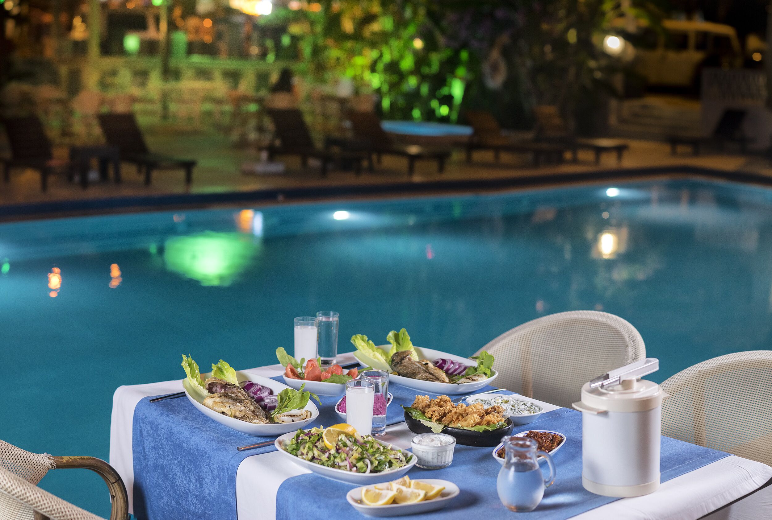 A dining table with various dishes and drinks by a poolside.