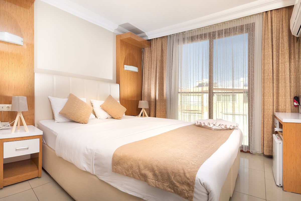 Habitación de hotel con una cama grande, mesitas de noche, un escritorio con silla y una ventana con cortinas.