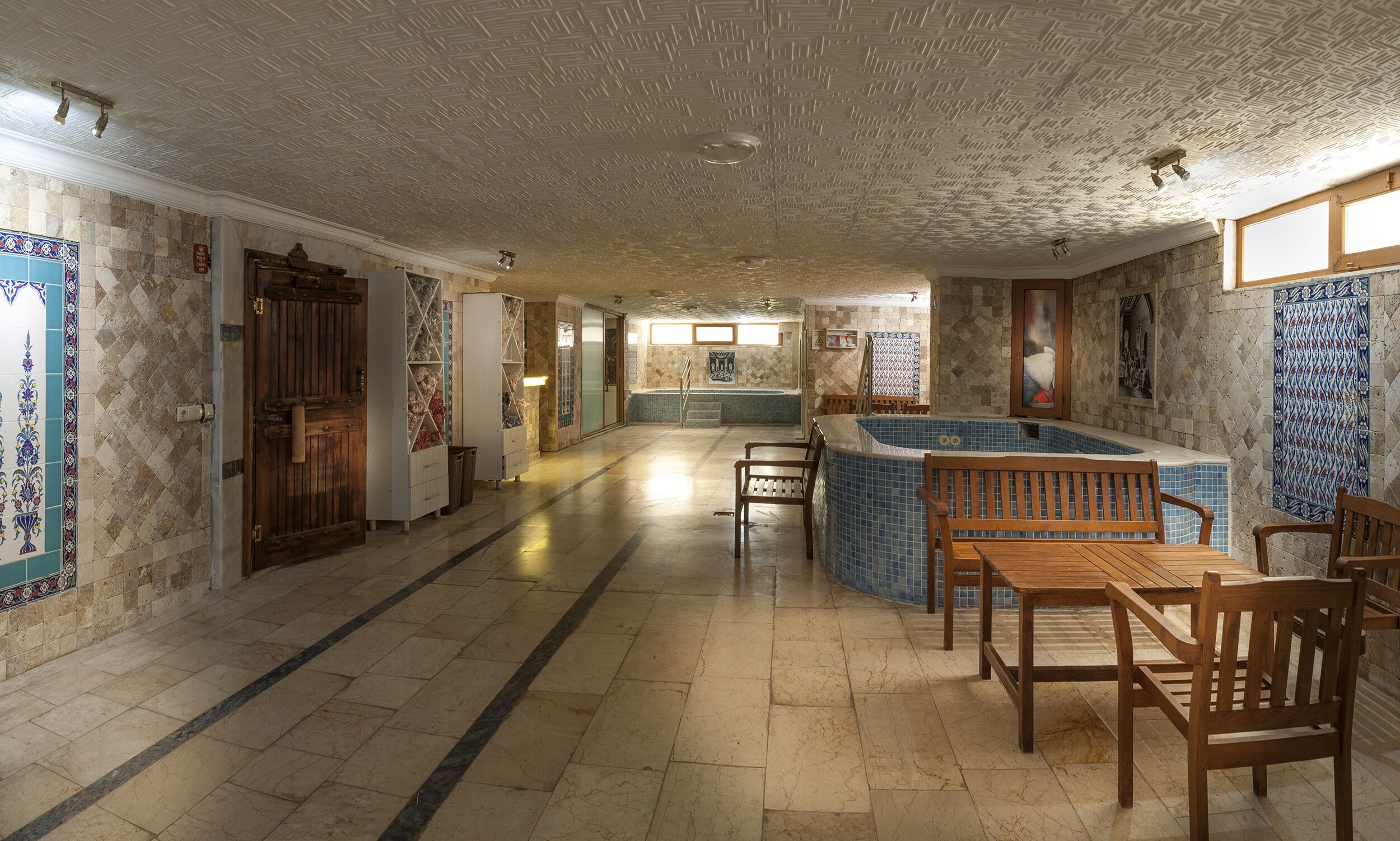 A spacious indoor area with a pool, wooden furniture, and tiled walls.