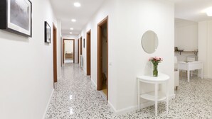 Un pasillo con paredes blancas, piso pulido con motas, cuadros enmarcados y un espejo redondo sobre un pedestal blanco.
