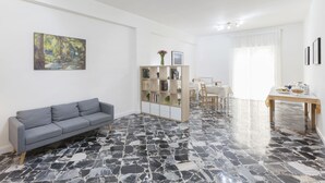 Un salón con un sofá gris, una estantería, una zona de comedor con una mesa y sillas, y un suelo de mármol.