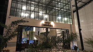 Un edificio moderno con una amplia entrada de vidrio, un letrero sobre la puerta y plantas de interior alrededor del área de acceso.